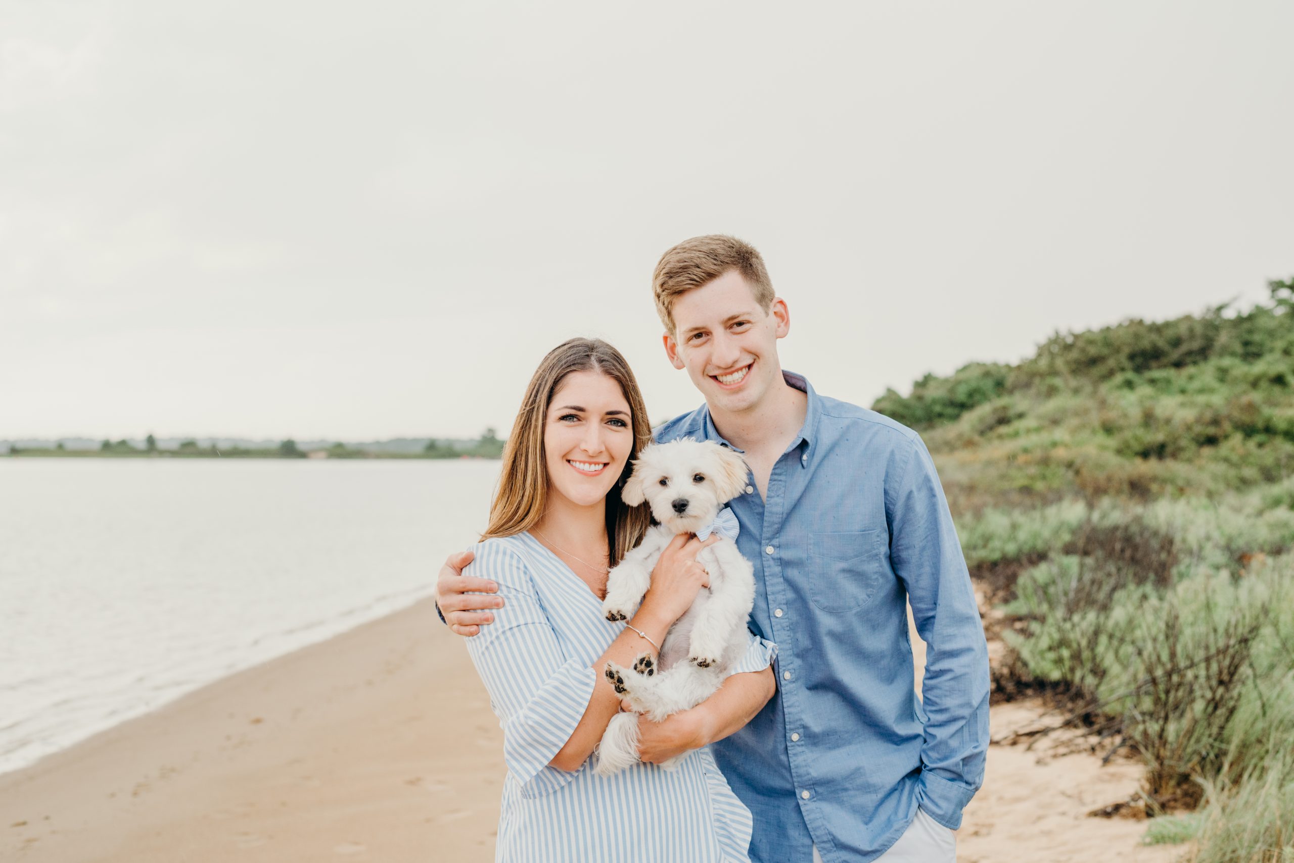 engaged couple smiling holding their dog on the beach during their Mashpee beach engagement session on cape cod massachusetts