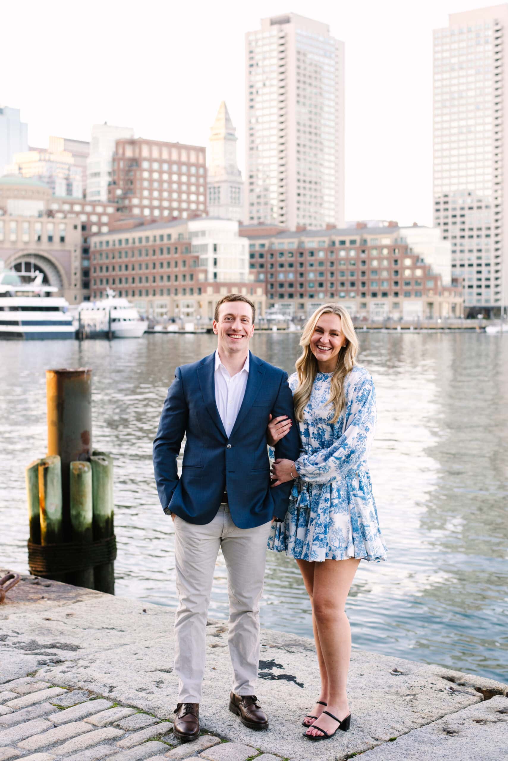 couple smiling in front of the boston skyline during their boston seaport engagement session captured by Meghan Lynch