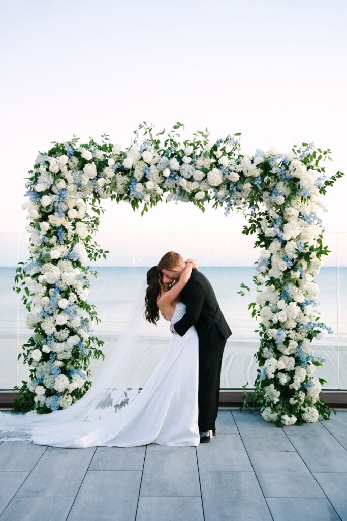 bride and groom sharing first kiss in front of a large hydrangea floral arch during their wychmere beach club dune wedding ceremony on Cape Cod 