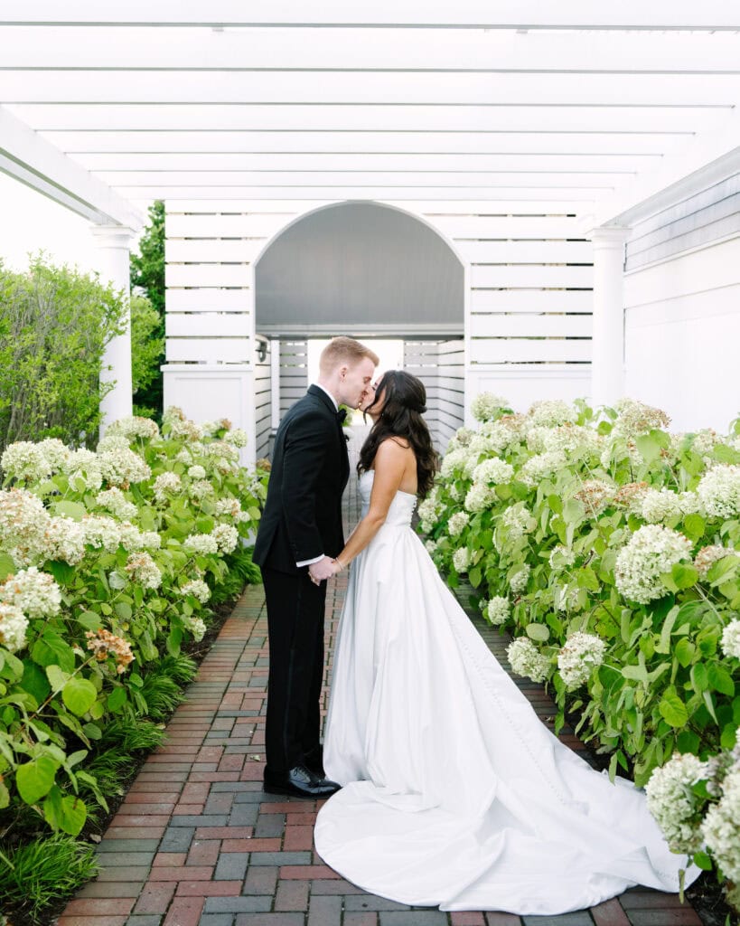 bride and groom kissing in between two rows of hydrangeas during their wychmere beach club wedding on cape cod