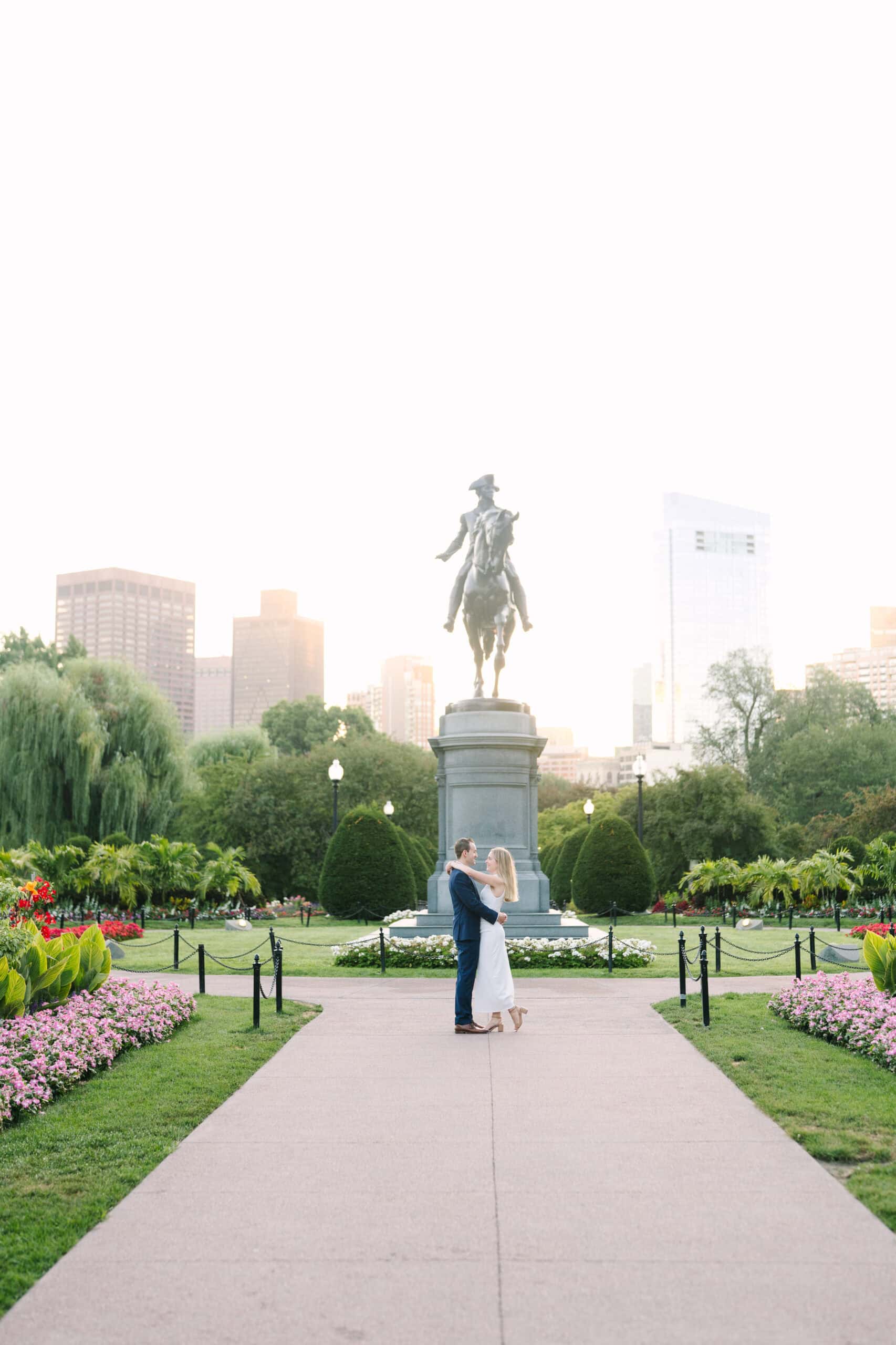 couple with arms wrapped around each other at sunrise in front of the george washington statue captured by boston engagement photographer meghan lynch
