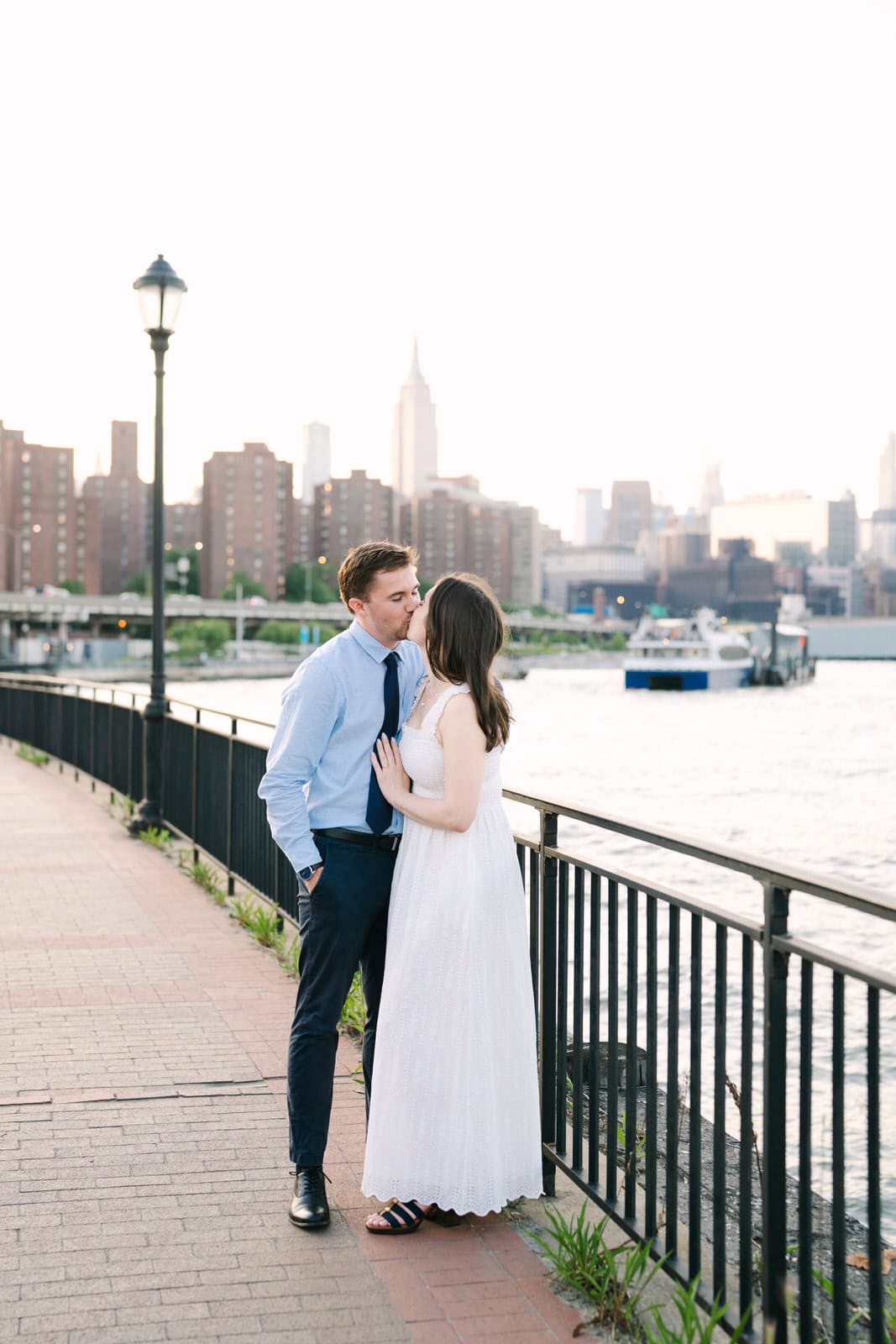 couples kissing by the east river with the empire state building behind during their nyc engagement session in stuytown
