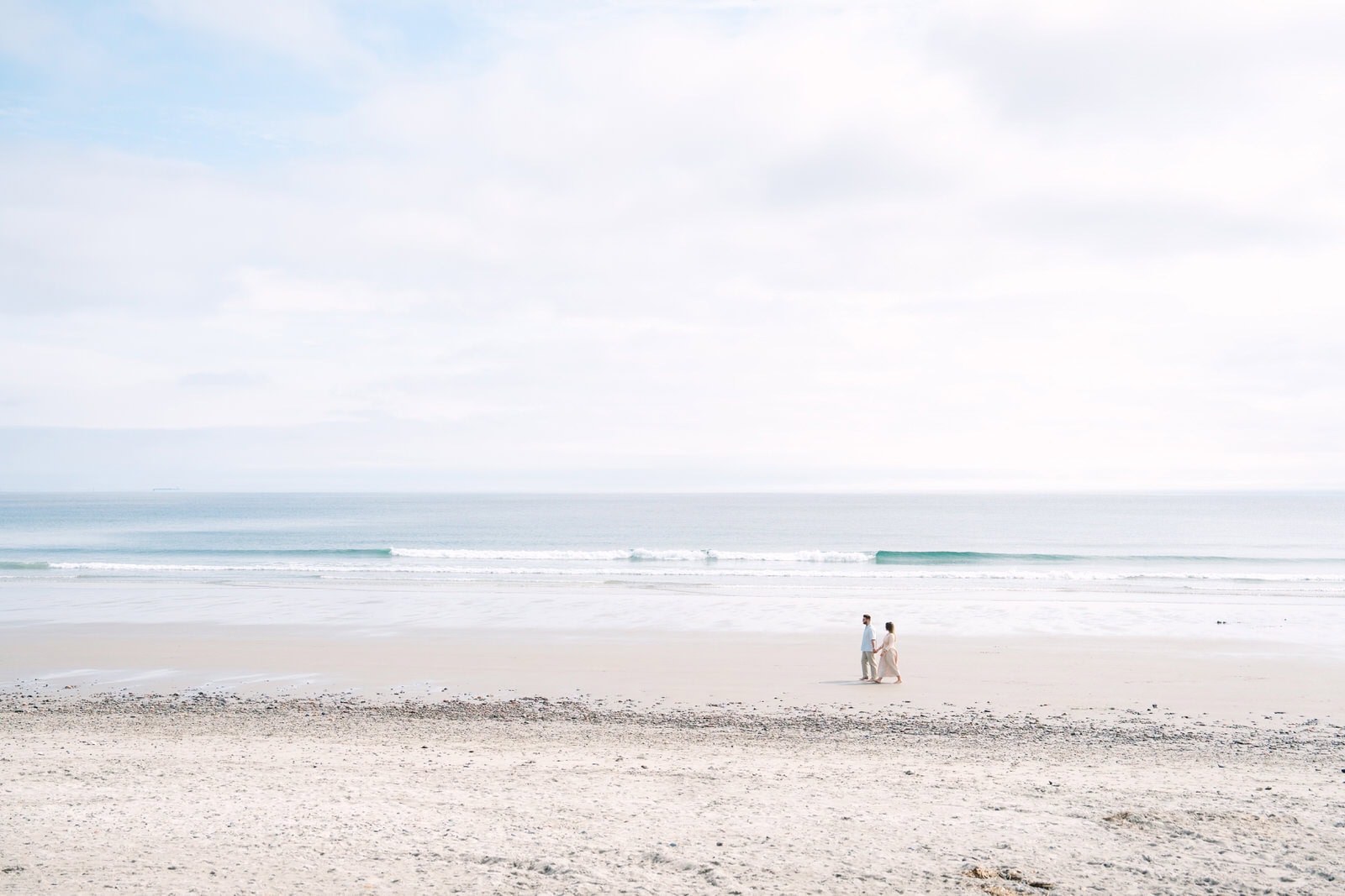 far away landscape shop of an engaged couple walking nantasket beach with the ocean behind them