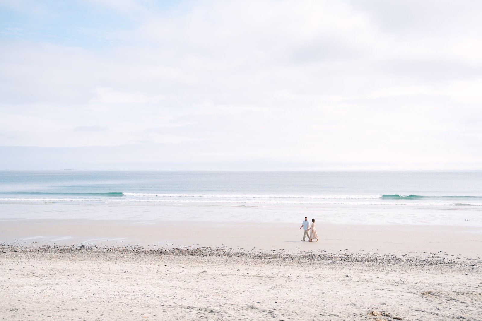 far away landscape shop of an engaged couple walking nantasket beach with the ocean behind them