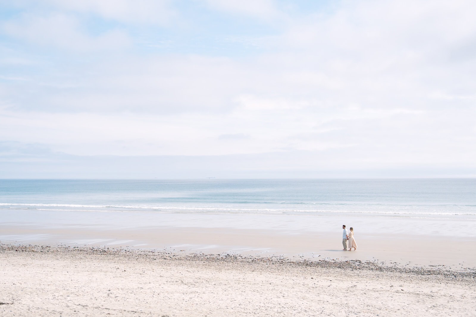 far away landscape shop of an engaged couple walking nantasket beach with the ocean behind them
