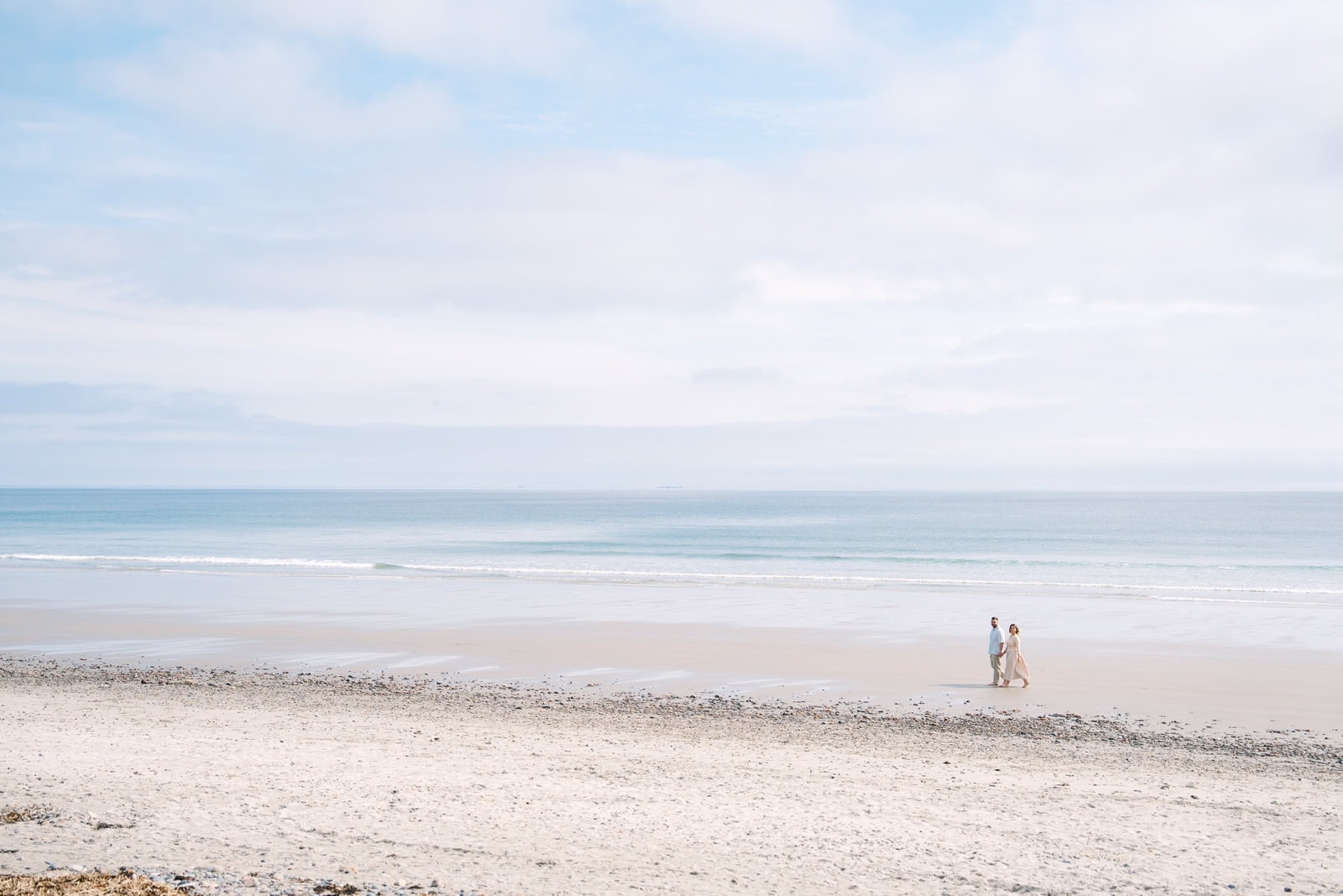 far away landscape shop of an engaged couple walking nantasket beach with the ocean behind them