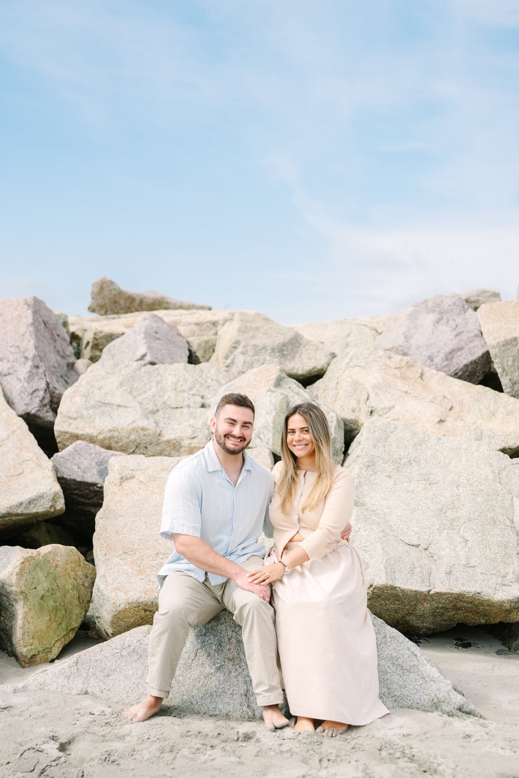 engaged couple sitting on rocks smiling at the camera during their nantasket beach engagement session