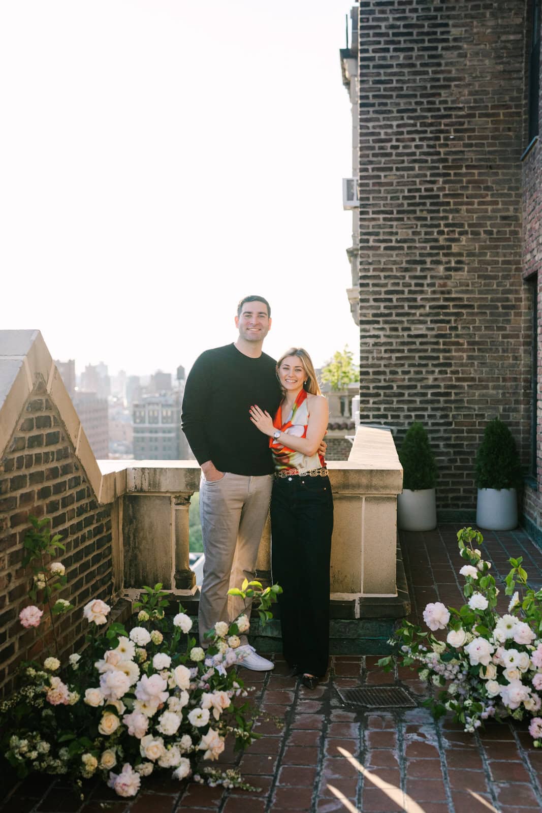 engaged couple smiling at the camera with two floral arrangements by them on a rooftop in greenwich village for their nyc surprise proposal session